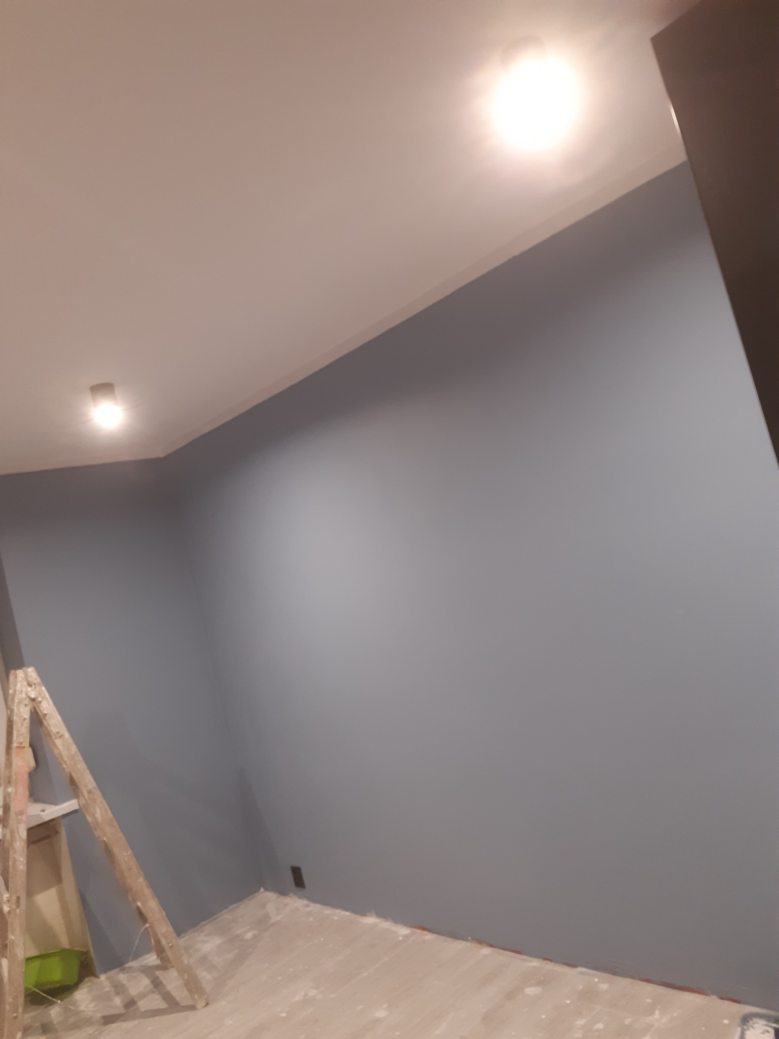 Świeżo pomalowane ściany na szaro w pokoju, widoczna drabina malarska i ślady farby na podłodze, oświetlenie punktowe na suficie.