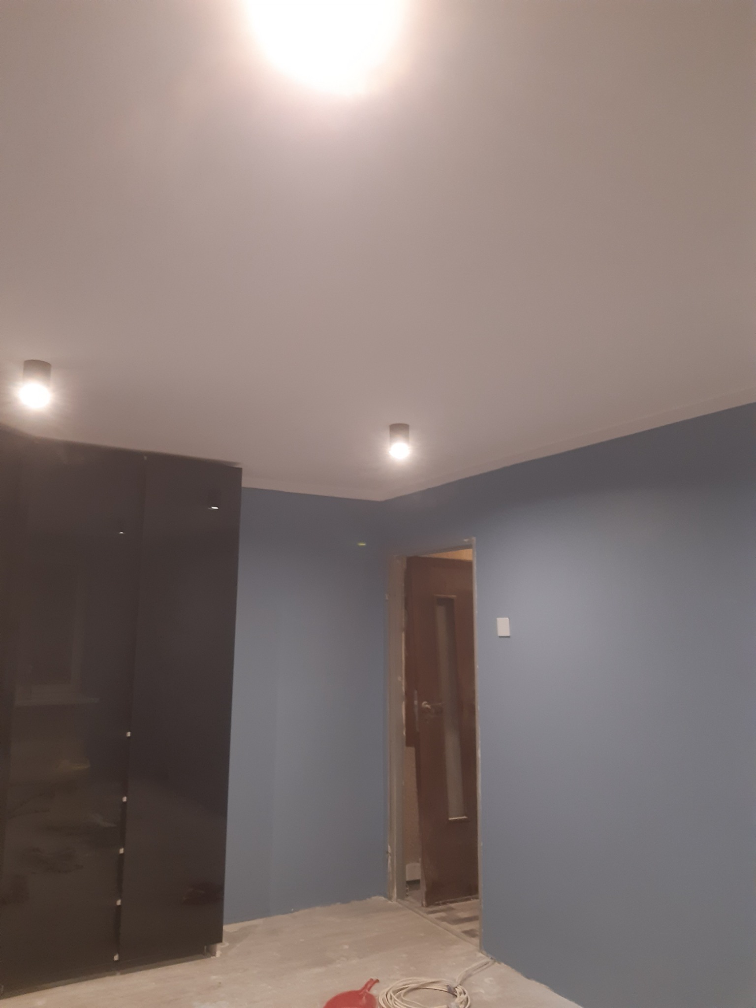 Pokój w trakcie malowania na szaro-niebieski kolor, widoczna czarna szafa z połyskiem, sufit z oświetleniem punktowym i wystające kable na podłodze.