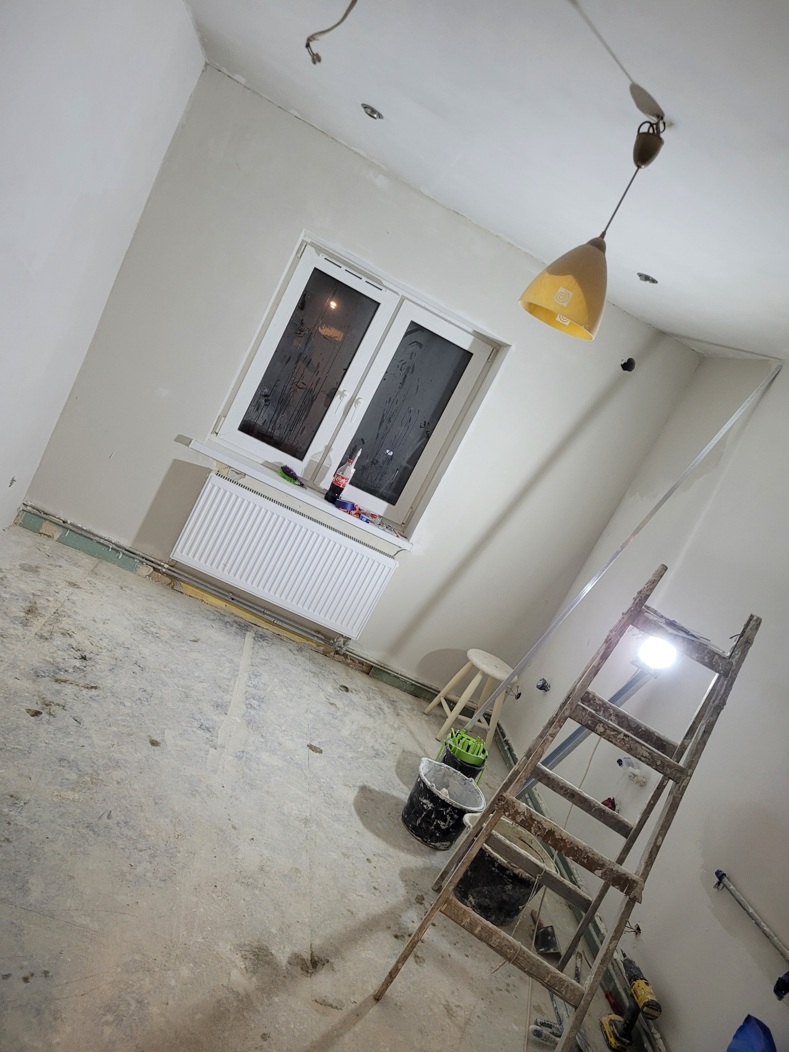 Pokój w trakcie remontu: ściany po szpachlowaniu, drabina z wiadrami farby, okno z grzejnikiem i napojem, wisząca lampa w tle