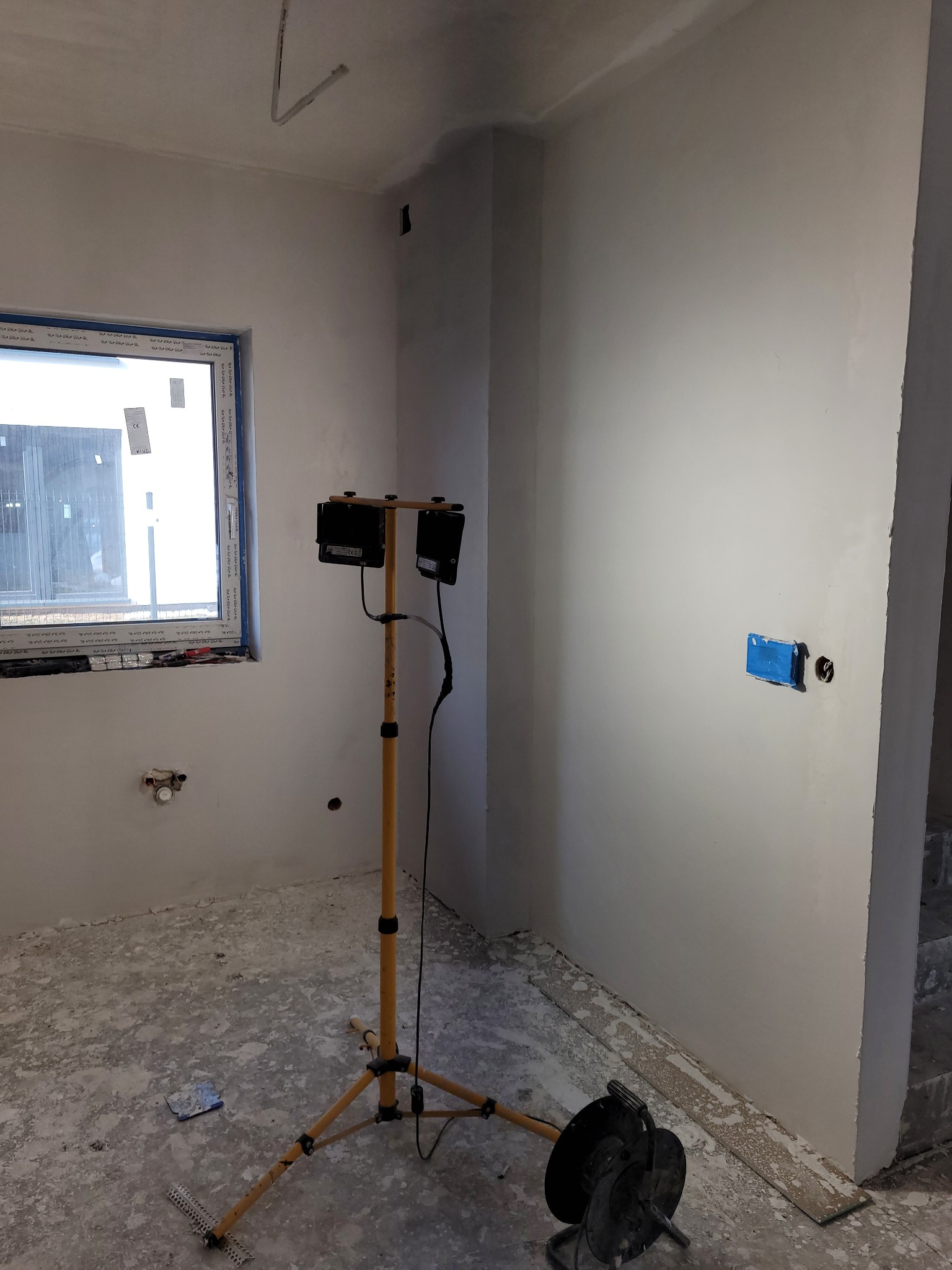 Pokój w trakcie remontu: świeżo pomalowane ściany, zaklejone gniazdko, lampa budowlana na statywie, resztki farby na podłodze i listwa przypodłogowa leżąca obok schodów.