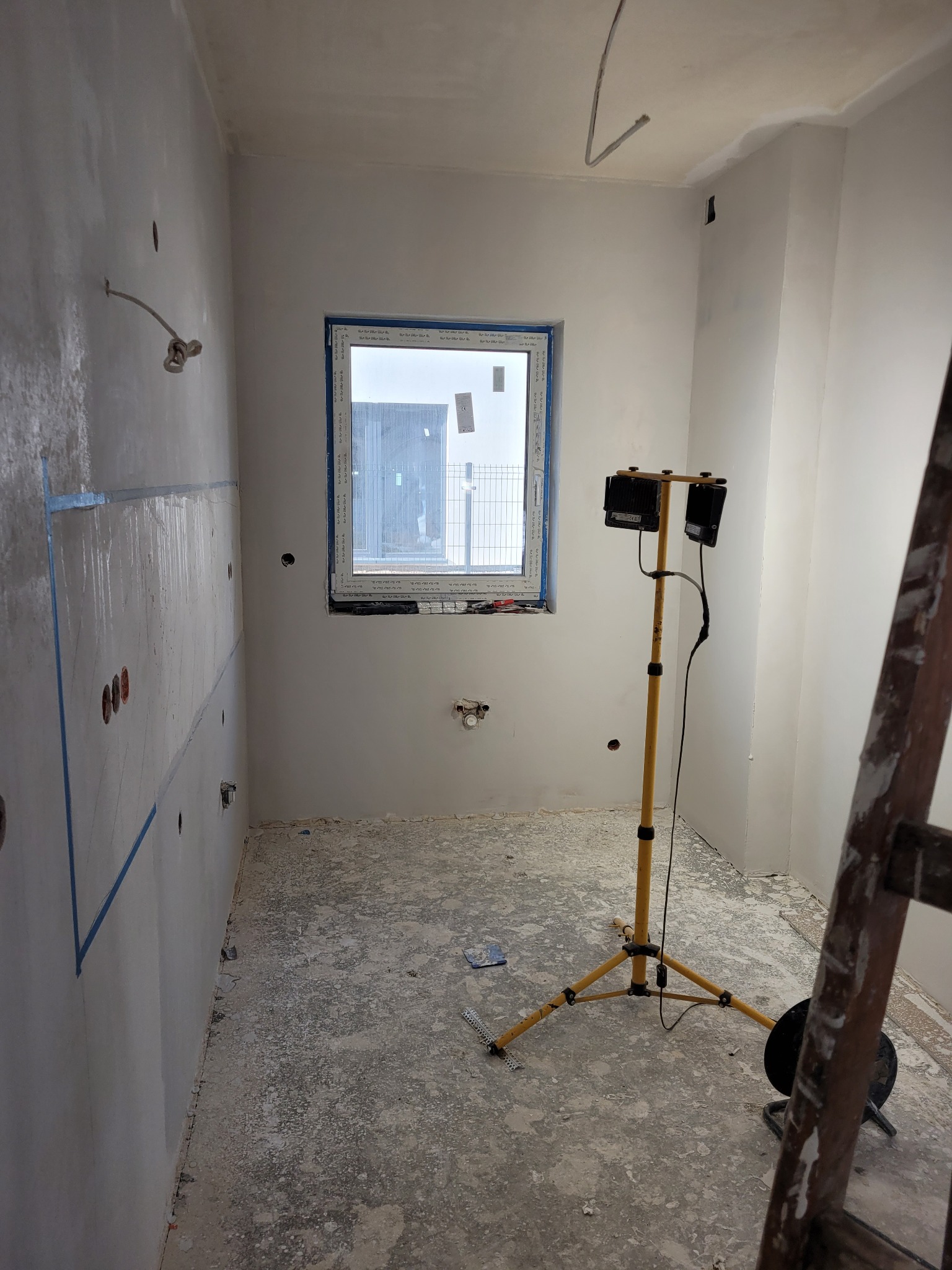 Wnętrze w trakcie prac przygotowawczych: ściany po gruntowaniu, zabezpieczone taśmą malarską, widoczne punkty elektryczne, okno z widokiem na zewnątrz, oświetlenie robocze na statywie i fragment...