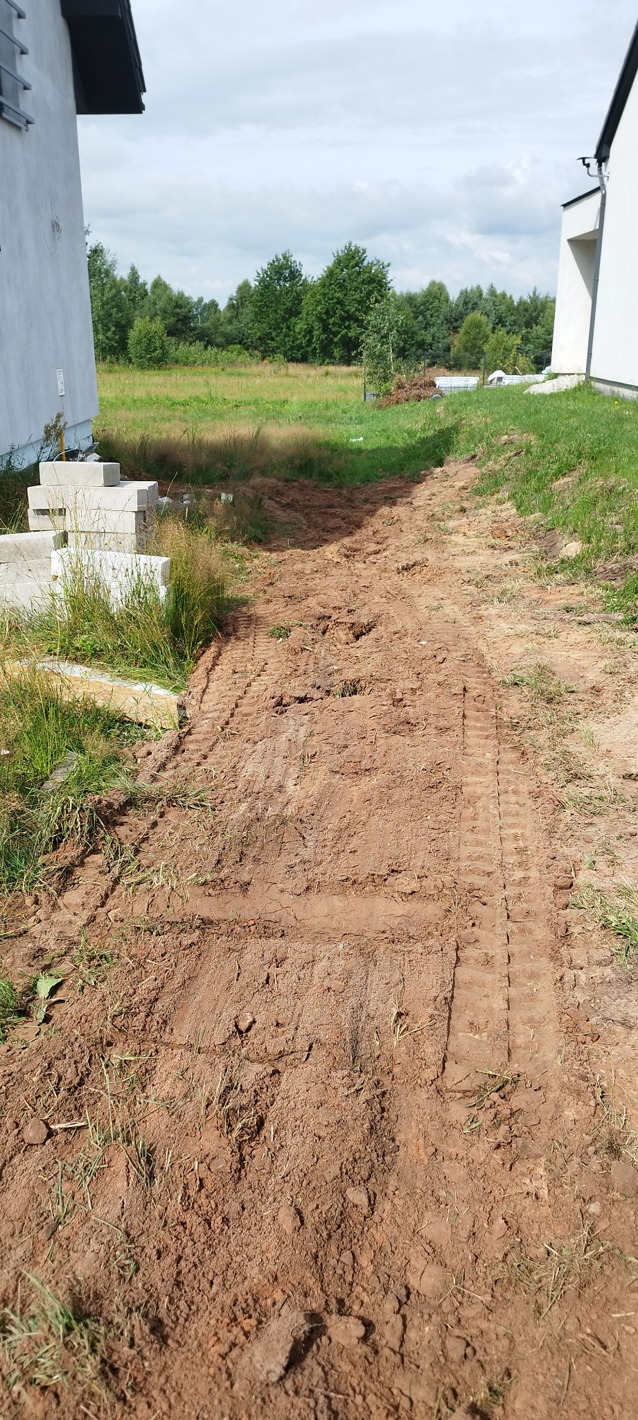 Przygotowanie terenu pod budowę domu: wyrównana ziemia z widocznymi śladami opon maszyny budowlanej, domy w tle.