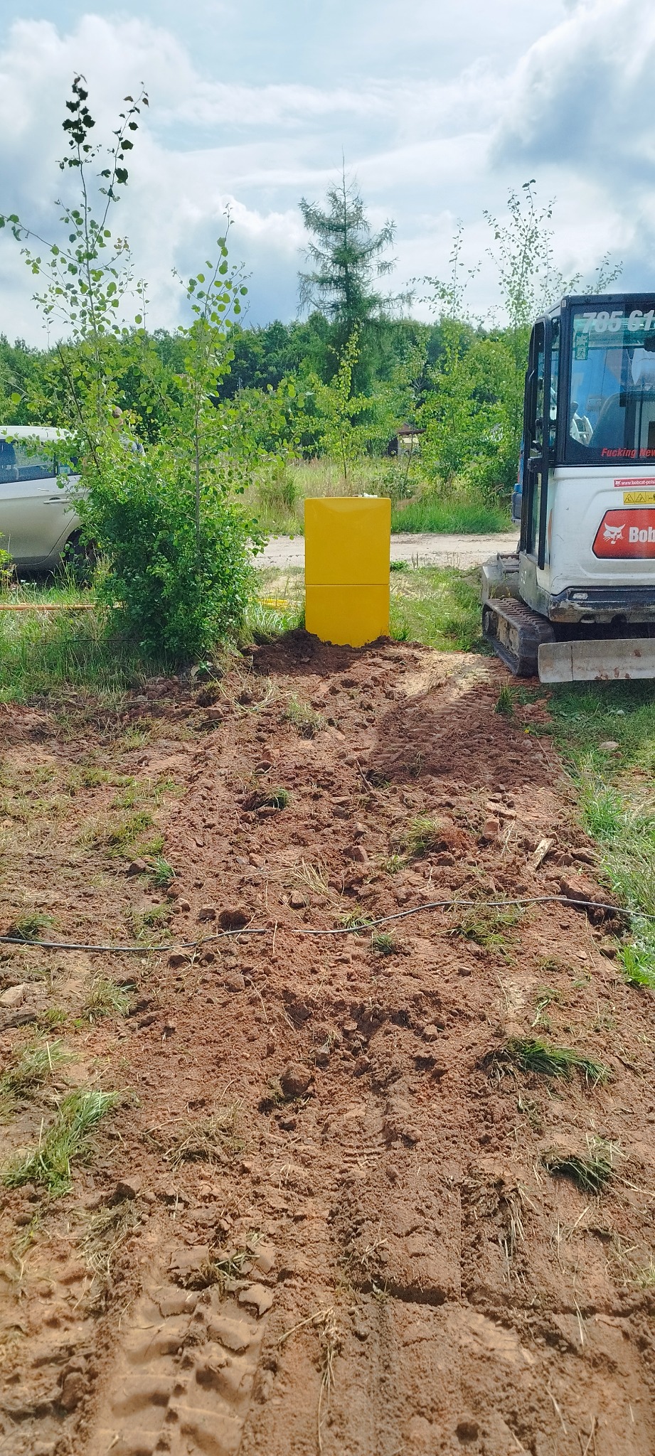 Żółta skrzynka z dwoma modułami ustawiona na ziemi obok małej koparki Bobcat z widocznymi śladami opon na ziemi i zielenią w tle.