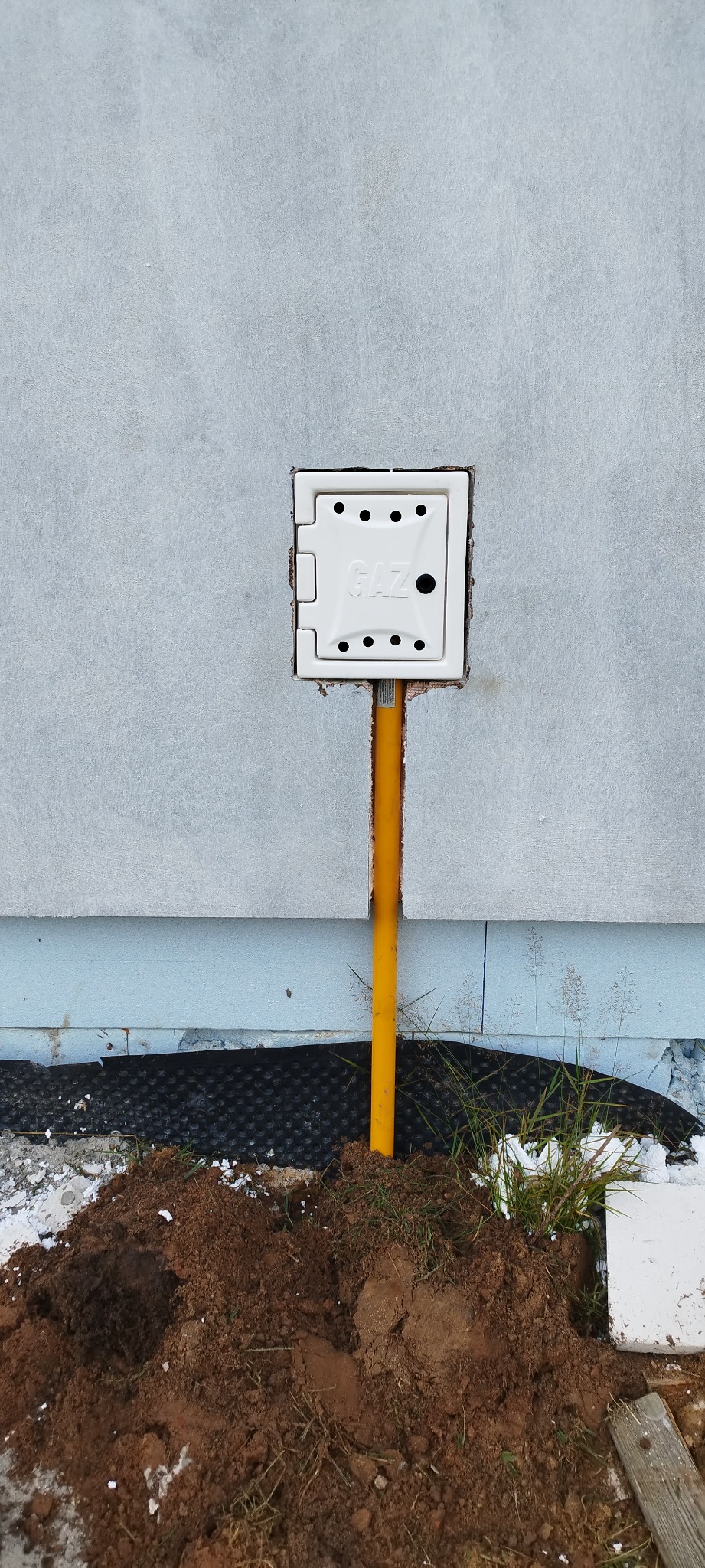 Biała skrzynka gazowa z żółtą rurą na tle szarej ściany budynku z widoczną warstwą izolacji i ziemią u podstawy.