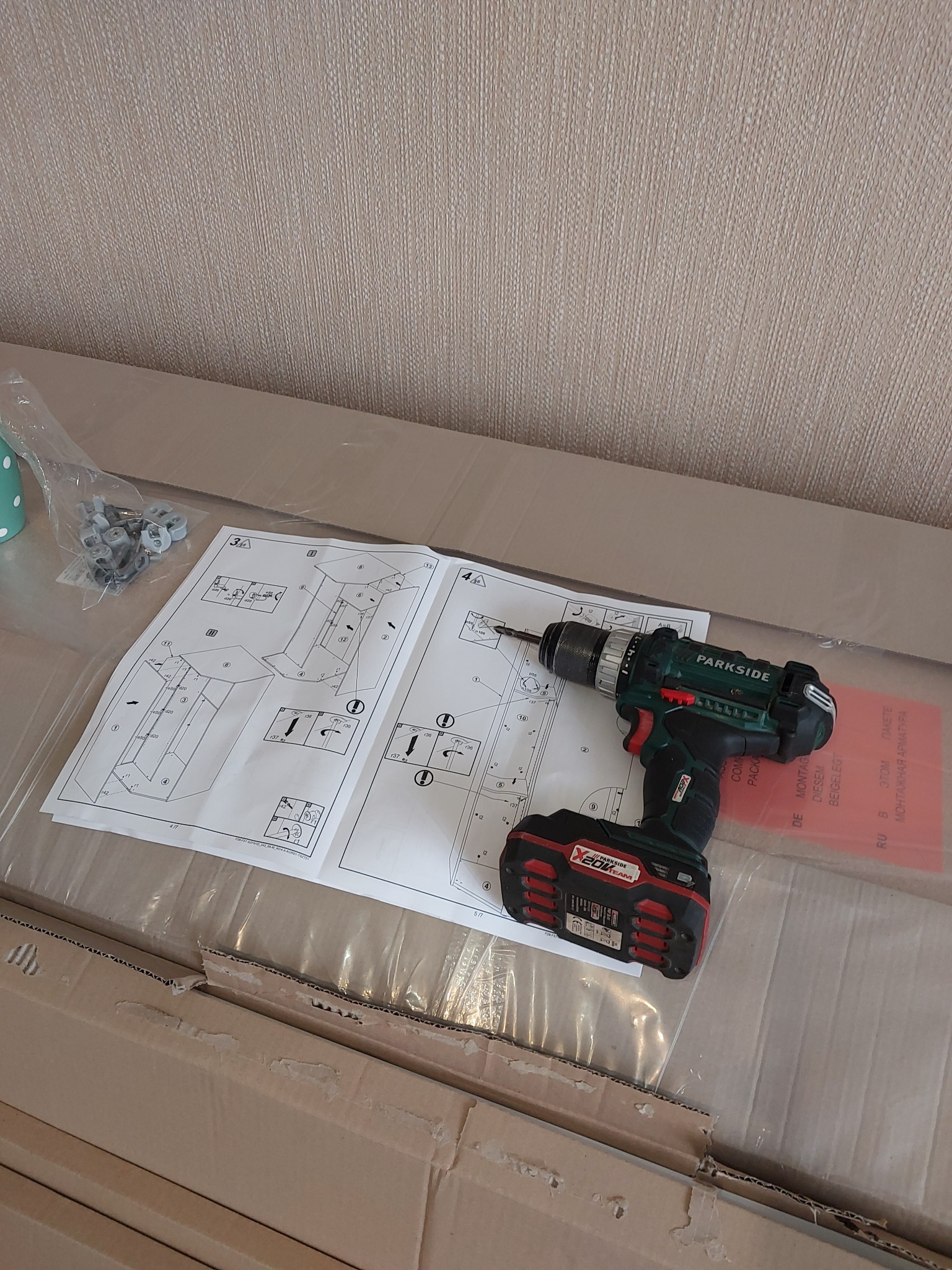 Akumulatorowa wiertarko-wkrętarka Parkside leży na instrukcji montażu mebli, obok woreczka z elementami montażowymi, całość na kartonowym opakowaniu.