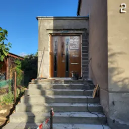 Nowe betonowe schody wejściowe do domu w trakcie budowy, z widocznymi narzędziami i materiałami budowlanymi oraz z częściowo zamontowanymi drzwiami wejściowymi.