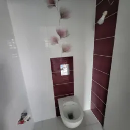 Biała toaleta wisząca w łazience z białymi i bordowymi płytkami, z motywem kwiatowym na ścianie i niedokończonym oświetleniem.