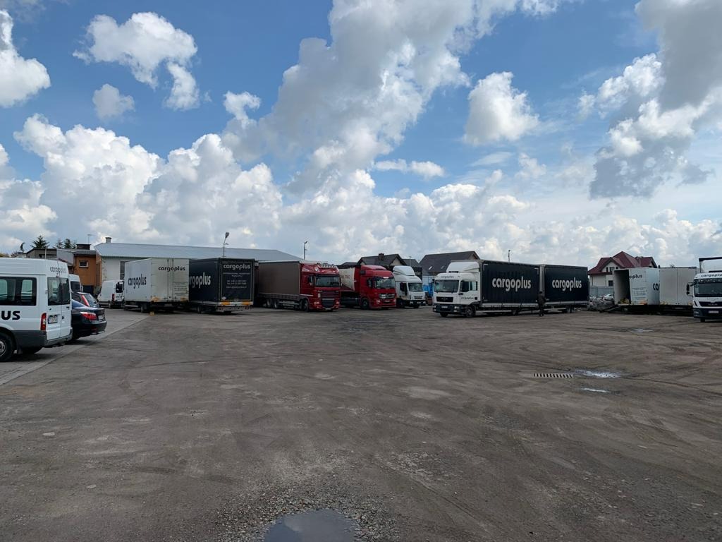 Flota ciężarówek z naczepami i busów firmy Cargoplus zaparkowana na placu, widoczne różne modele i kolory pojazdów, na naczepach logo firmy, w tle budynki i błękitne niebo z chmurami.