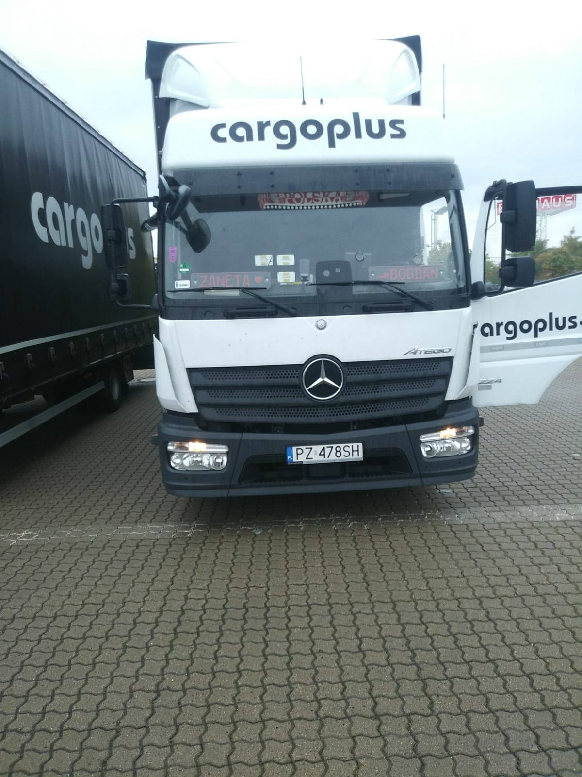 Ciężarówka Mercedes Atego firmy Cargoplus na parkingu z kostki brukowej, widoczna tablica rejestracyjna PZ 478SH oraz naklejki z imionami 'Żaneta' i 'Bogdan' na przedniej szybie.