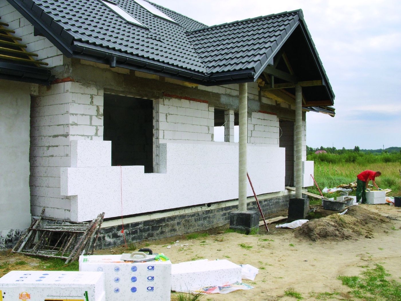 Ocieplanie ściany zewnętrznej budynku styropianem, widoczny pracownik w czerwonej koszulce i zielonych spodniach, białe bloki styropianu oraz elementy konstrukcyjne dachu z czarną dachówką.