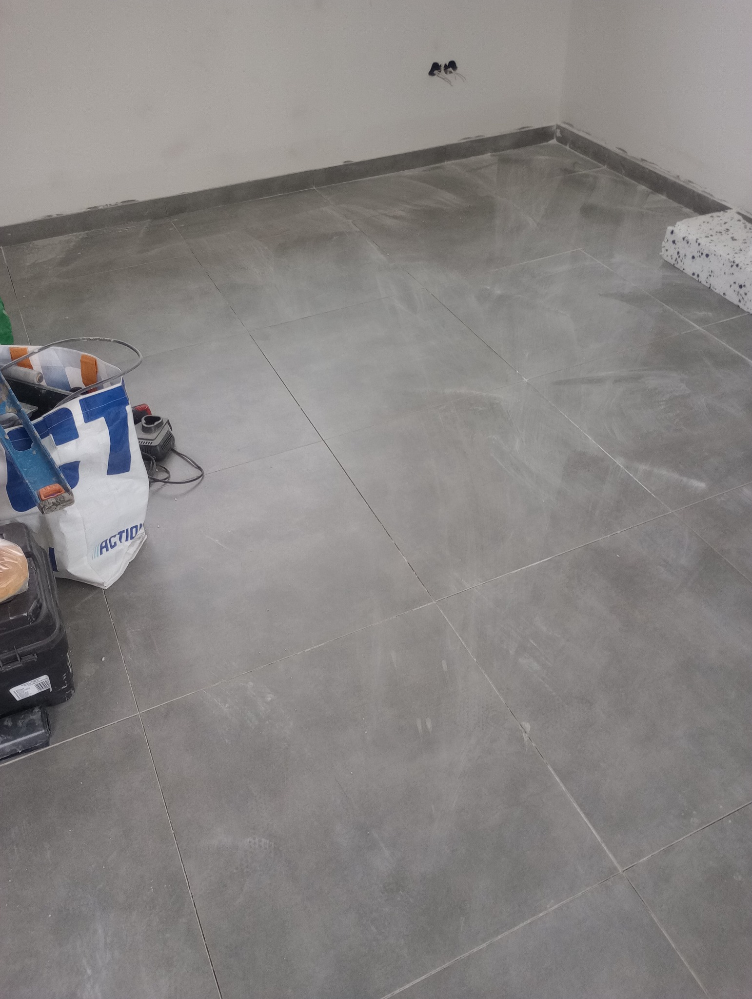 Ułożone szare płytki podłogowe w trakcie prac wykończeniowych, widoczne narzędzia i torba budowlana w rogu pomieszczenia, ślady fugi na powierzchni płytek.