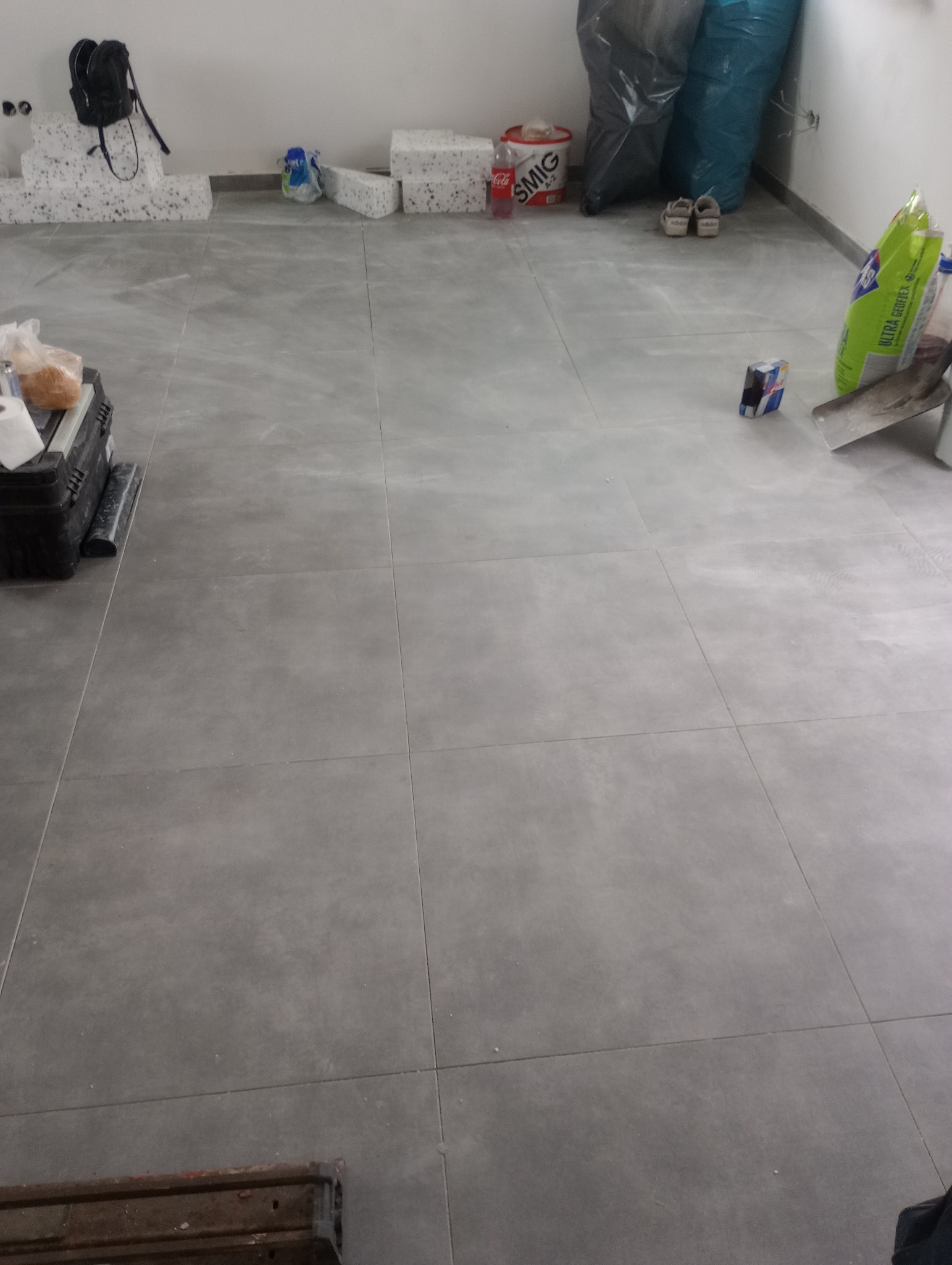 Szare płytki podłogowe ułożone w kwadraty, widoczne materiały budowlane, torba sportowa i narzędzia, sugerujące etap prac wykończeniowych w pomieszczeniu.