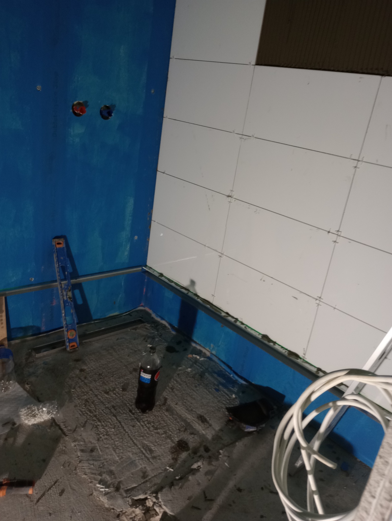 Niedokończona łazienka z niebieskimi ścianami, częściowo wyłożona białymi płytkami, poziomica oparta o ścianę, butelka napoju na podłodze, widoczne kable i instalacja wodna.
