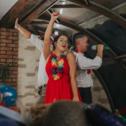 Rozbawiona kobieta w czerwonej sukience i kwiatowym naszyjniku, z podniesioną ręką, śpiewa do mikrofonu podczas imprezy, w tle mężczyzna trzyma się drewnianej belki stropowej, a drugi śpiewa...