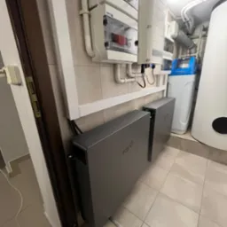 Dwa szare moduły magazynu energii Fox ESS zamontowane w pomieszczeniu z płytkami, widoczne także białe skrzynki elektryczne na ścianie i system uzdatniania wody w tle.