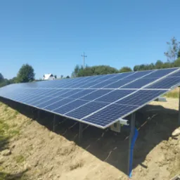 Długa instalacja fotowoltaiczna na zboczu z widocznymi niebieskimi rurami osłonowymi kabli, w tle dom i drzewa pod błękitnym niebem.