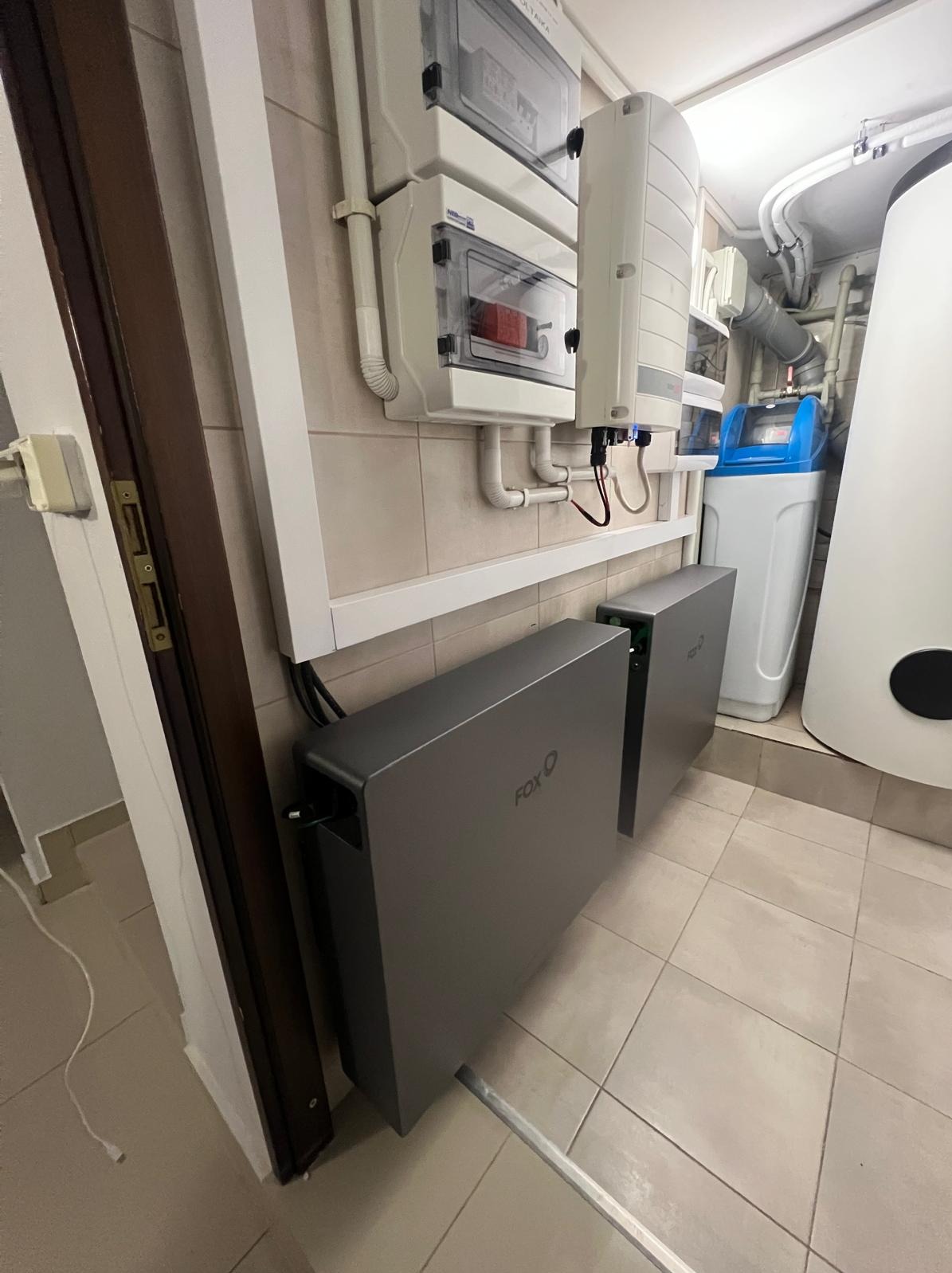 Dwa szare moduły magazynu energii Fox ESS zamontowane w pomieszczeniu z płytkami, widoczne także białe skrzynki elektryczne na ścianie i system uzdatniania wody w tle.