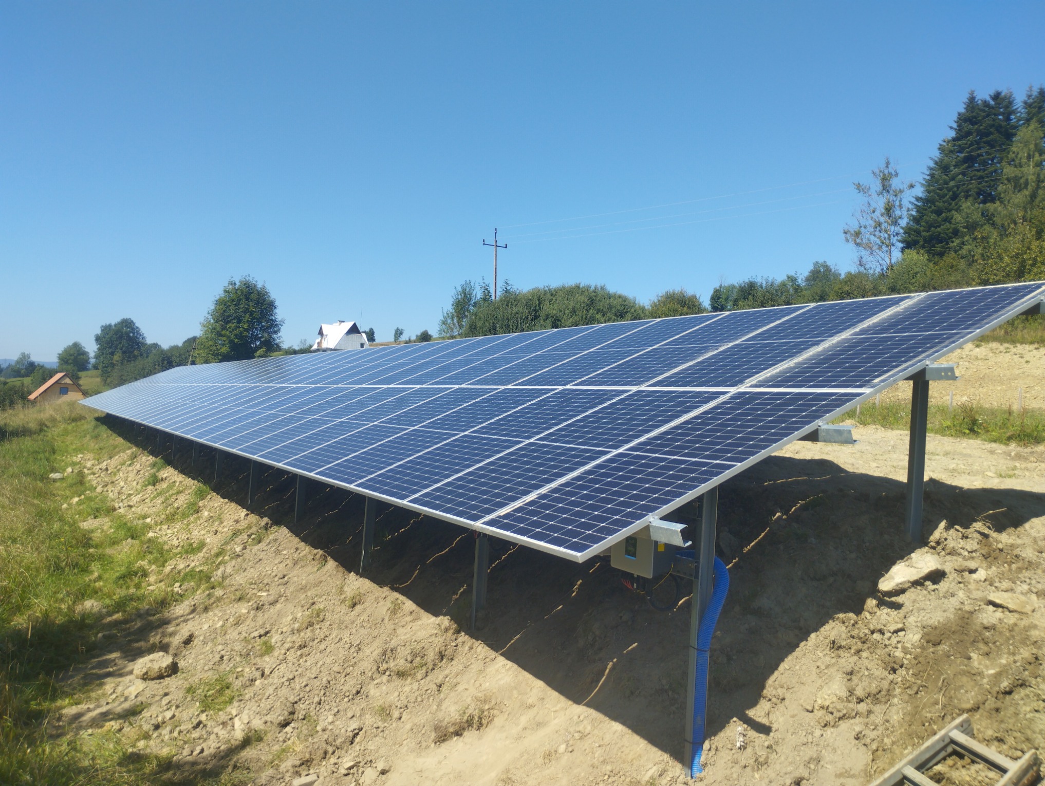 Długa instalacja fotowoltaiczna na zboczu z widocznymi niebieskimi rurami osłonowymi kabli, w tle dom i drzewa pod błękitnym niebem.