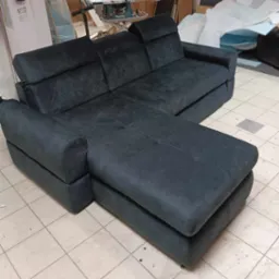 Ciemnoszara, rozkładana sofa narożna z regulowanymi zagłówkami, widoczna w warsztacie na kafelkowanej podłodze, z narzędziami i materiałami w tle.