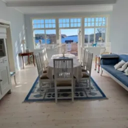 Jasny, przestronny salon z widokiem na morze, wyposażony w białe meble w stylu skandynawskim, duży stół jadalny z krzesłami i niebieską sofę z poduszkami, doświetlony naturalnym światłem.