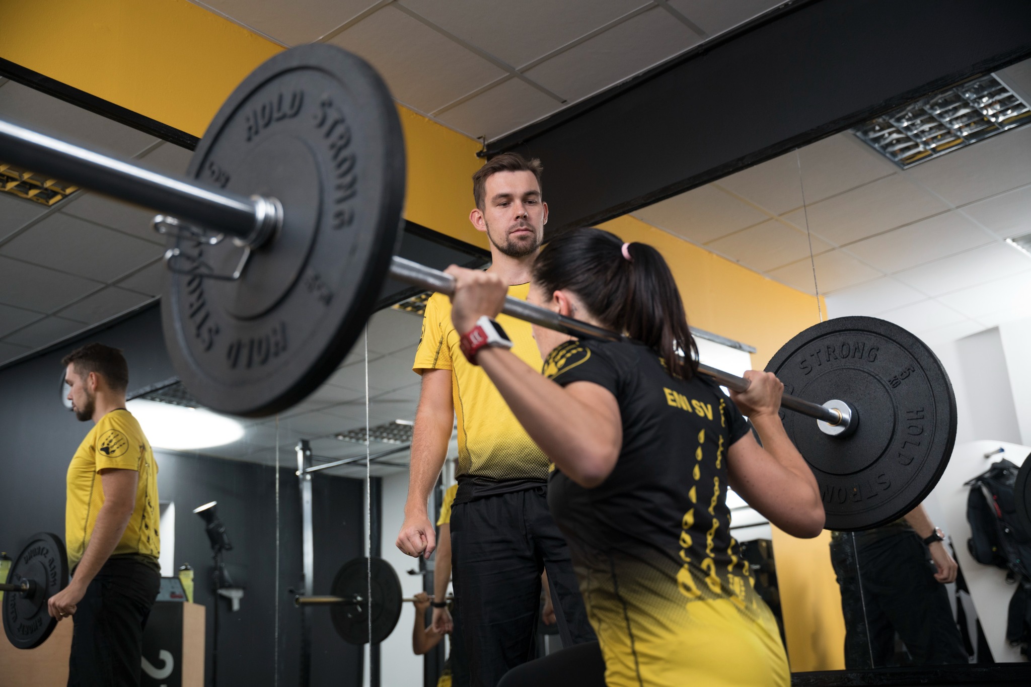 Kobieta wykonuje przysiad ze sztangą pod okiem trenera w siłowni z lustrami.