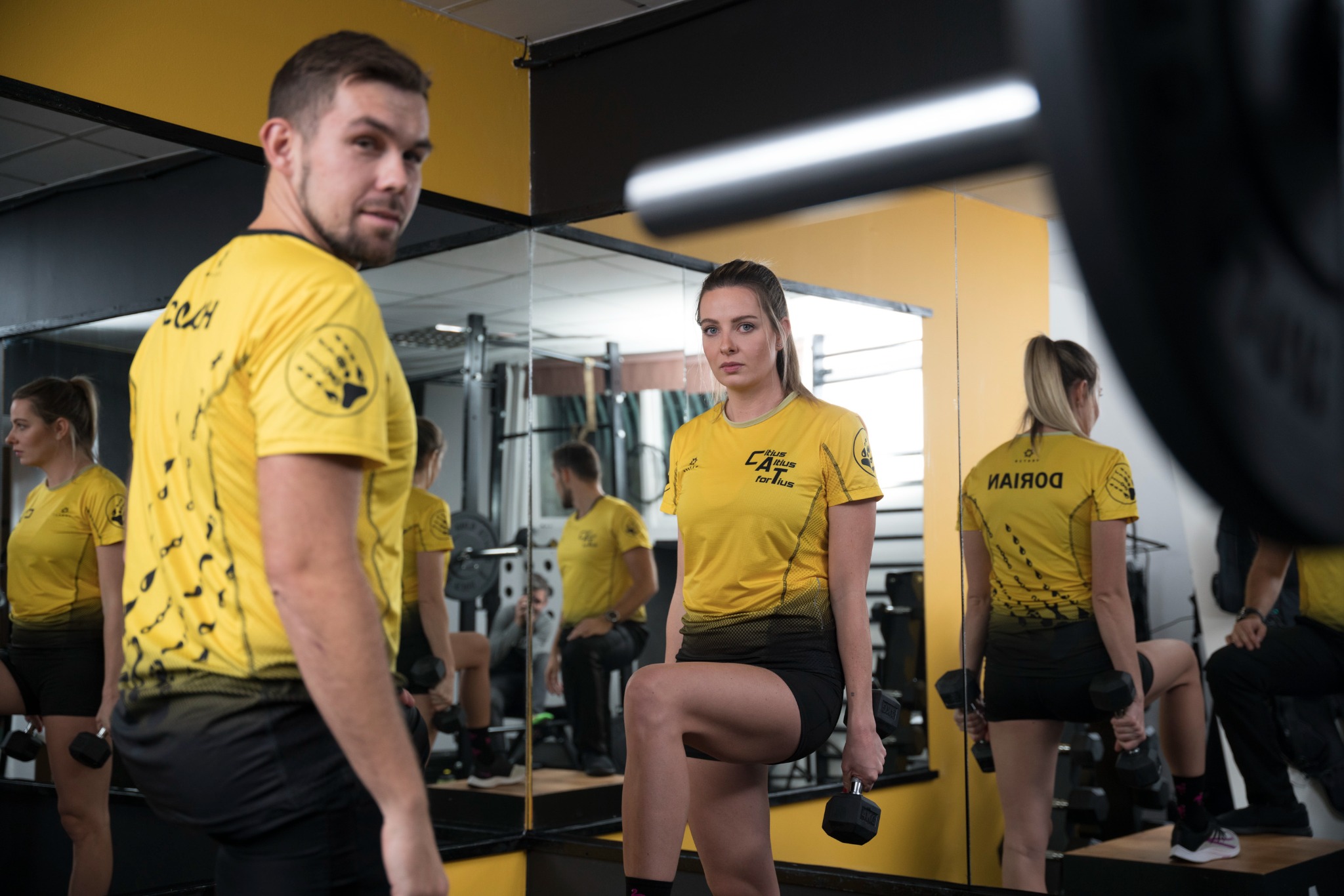 Para w żółtych koszulkach sportowych ćwiczy z hantlami w siłowni we Wrocławiu, widoczny napis COACH na koszulce mężczyzny oraz logo firmy na koszulce kobiety.