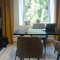 Biuro z czarnym biurkiem, laptopem, krzesłami z brązowej tkaniny i oknem z widokiem na drzewa, oświetlone żółtymi zasłonami.