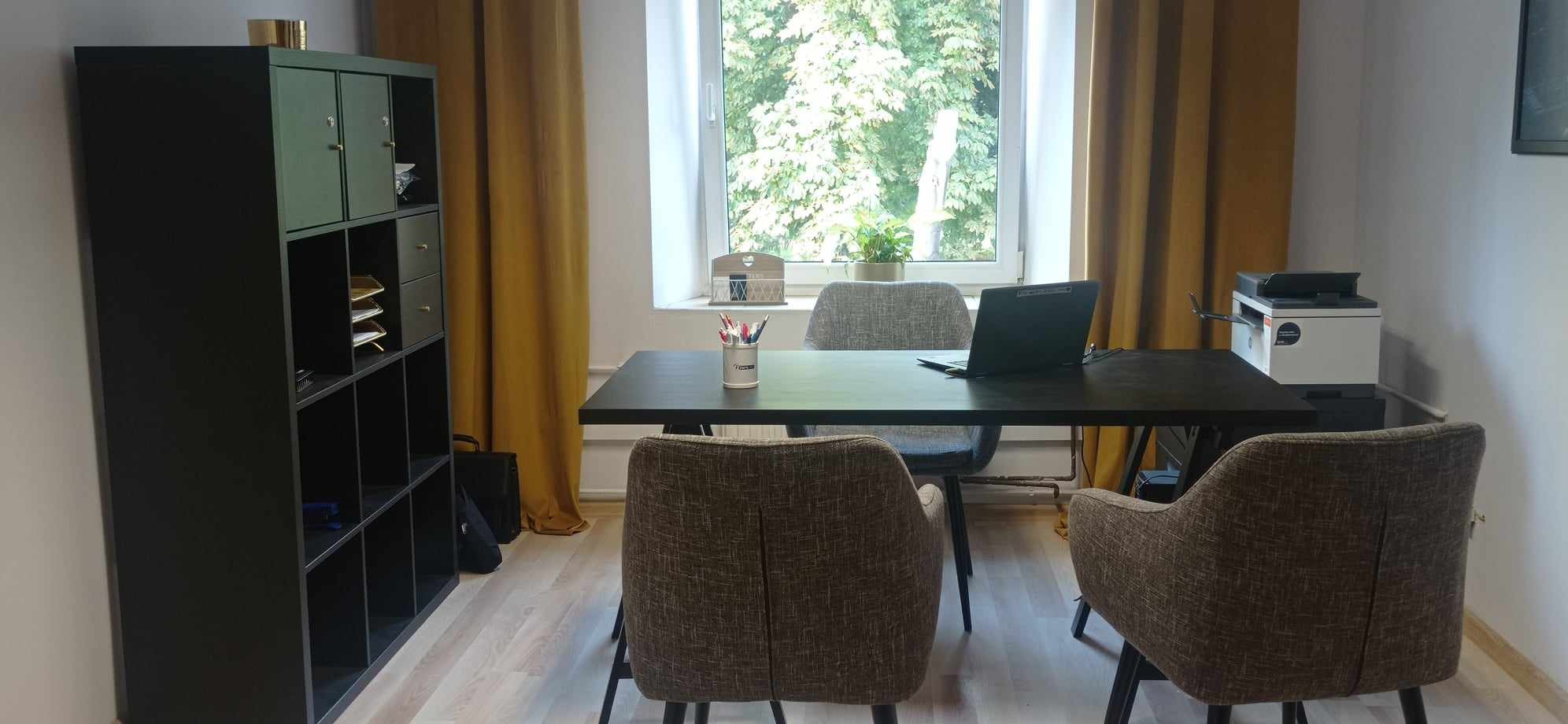 Biuro z czarnym biurkiem, laptopem, krzesłami z brązowej tkaniny i oknem z widokiem na drzewa, oświetlone żółtymi zasłonami.