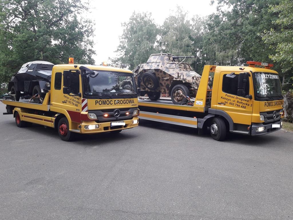 Dwie żółte lawety 'Pomoc Drogowa' przewożące pojazdy: czarny, sportowy samochód i wojskowy pojazd opancerzony z kamuflażem.