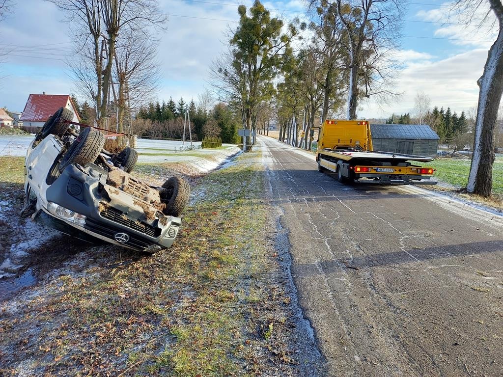 Przewrócony na bok biały bus w rowie, żółta laweta na drodze w tle, zimowy krajobraz.