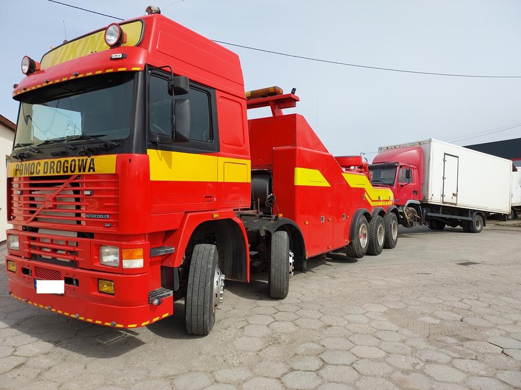 Czerwona ciężarówka pomocy drogowej Volvo z żółtymi pasami i napisem 'Pomoc Drogowa' holuje inną czerwoną ciężarówkę z białą naczepą na brukowanym placu.