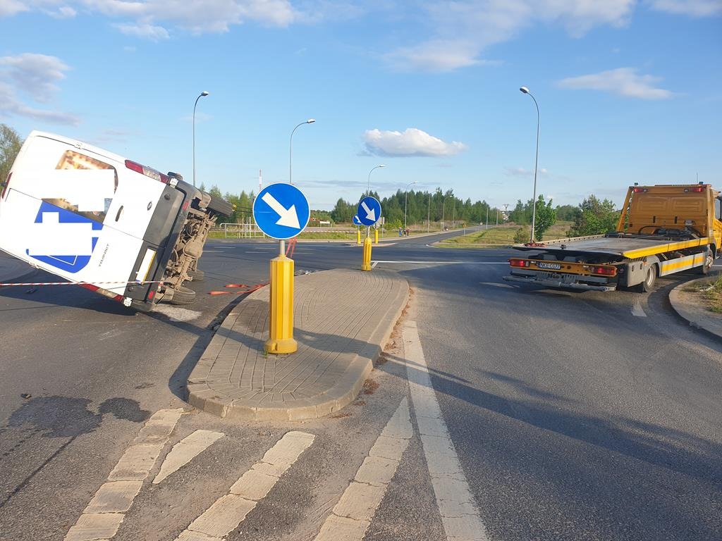 Przewrócony na bok biały bus na skrzyżowaniu, zabezpieczony taśmą, w tle żółta laweta i niebieskie znaki drogowe.