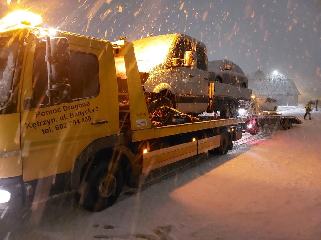Żółta ciężarówka pomocy drogowej z załadowanym samochodem osobowym podczas śnieżycy, widoczne napisy informacyjne na kabinie.