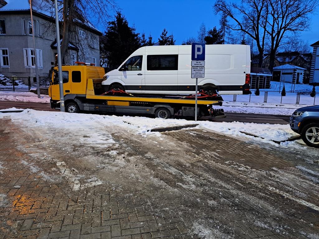 Żółta laweta z białym busem na platformie, zaparkowana w Kętrzynie w zimowej scenerii, widoczny znak parkingu.
