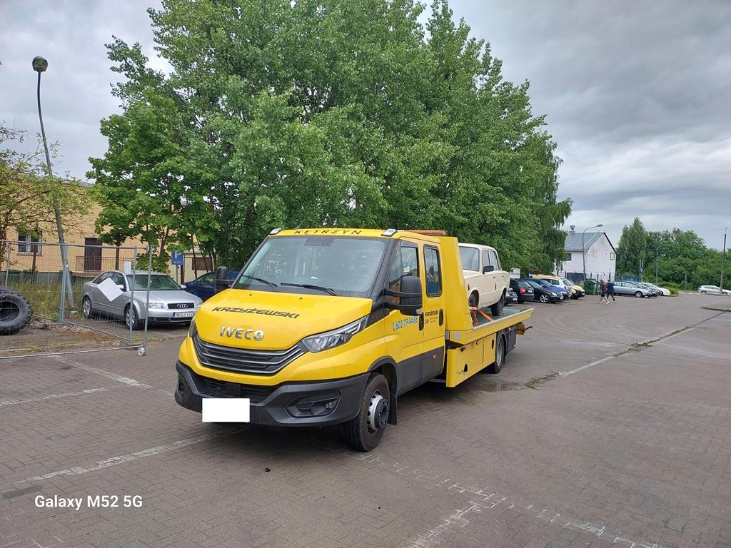 Żółta laweta Iveco z załadowanym białym samochodem osobowym, zaparkowana na placu z kostki, w tle zieleń drzew i budynki.