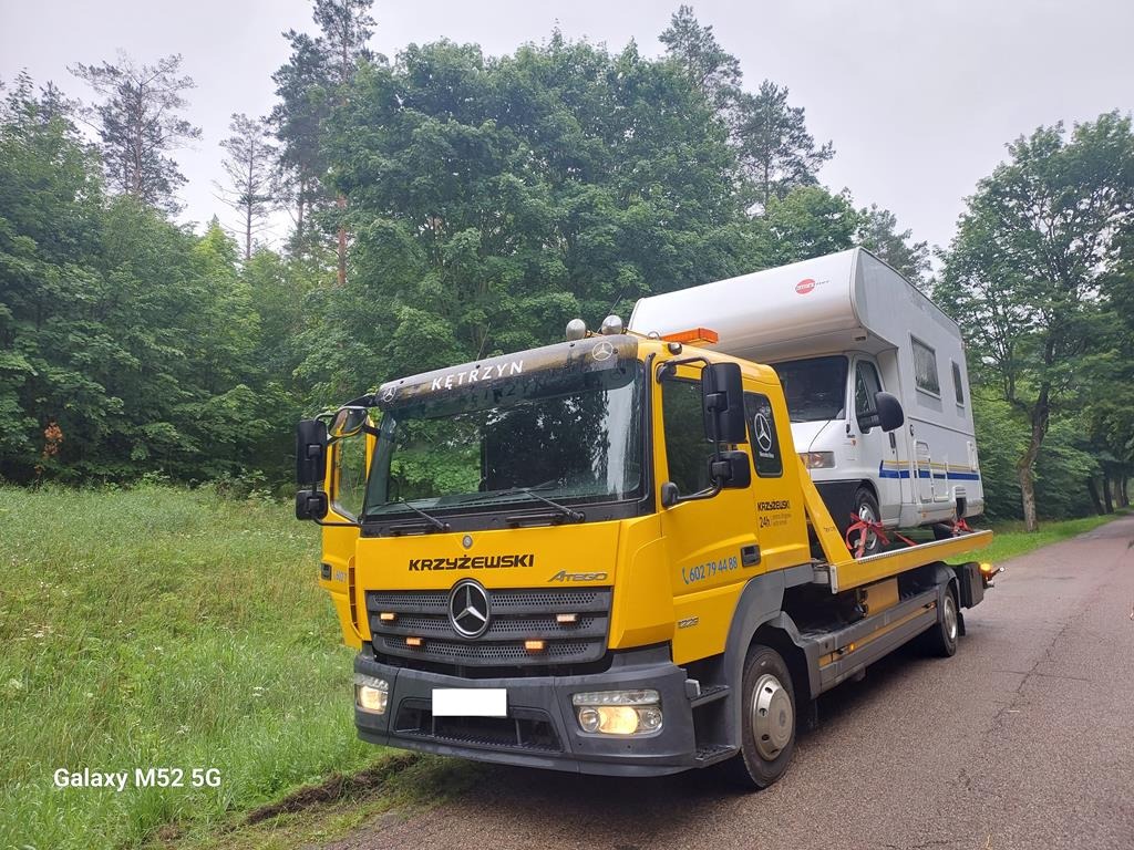 Żółta laweta Mercedes-Benz z napisem 'Kętrzyn' i 'Krzyżewski' przewozi biały kamper na tle zielonego lasu i drogi asfaltowej.