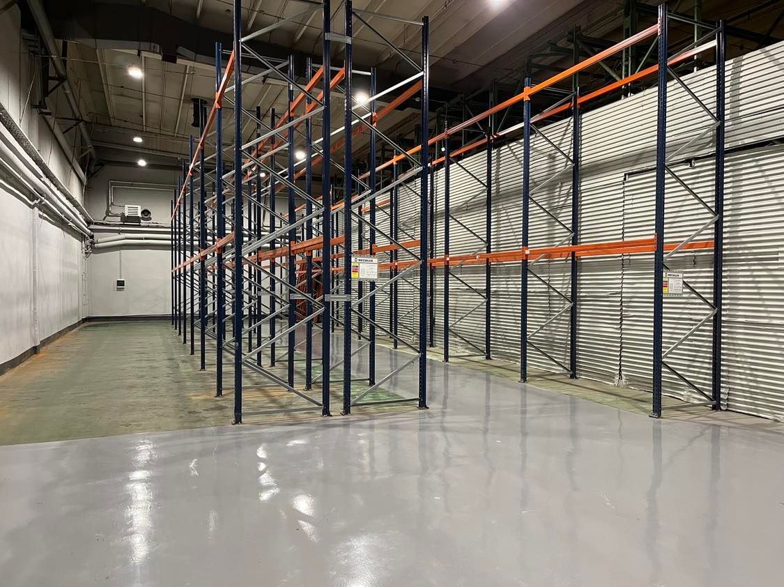 Puste, metalowe regały magazynowe z pomarańczowymi poprzeczkami w przestronnym, jasnym magazynie z szarą, lśniącą posadzką i blachą falistą na ścianie.