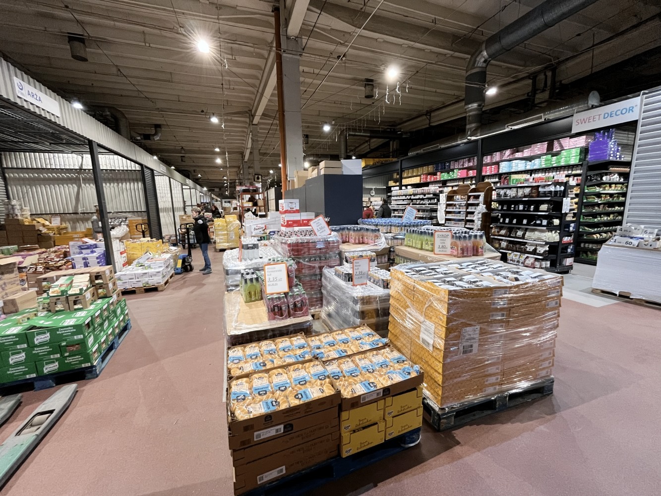 Wnętrze dużego magazynu hurtowego z paletami pełnymi zapakowanych produktów spożywczych, widoczne regały z towarem w tle, oświetlenie sufitowe.