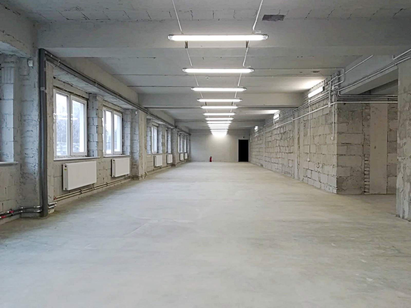 Puste, jasne pomieszczenie magazynowe z betonową podłogą, rzędem okien po lewej stronie i ścianami z bloczków betonowych po prawej, oświetlone lampami fluorescencyjnymi na suficie, z gaśnicą w oddali.