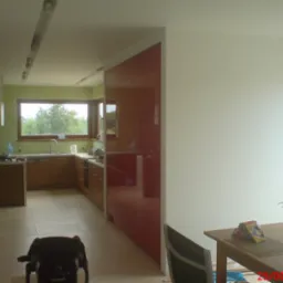 Jasna kuchnia w trakcie adaptacji, widoczne drewniane szafki, czerwony panel ścienny, okno z widokiem na zieleń oraz stół z bukietem kwiatów w rogu pomieszczenia.