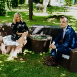 Kobieta w kwiecistej sukience i mężczyzna w granatowym garniturze siedzą na wiklinowych fotelach na trawie, z wiklinowym stolikiem i skrzynią w tle.