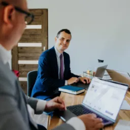 Spotkanie biznesowe: mężczyzna w garniturze z fioletowym krawatem uśmiecha się, siedząc przy drewnianym stole z laptopem, obok współpracownicy i kolegi w szarej marynarce, pracujących na swoich...