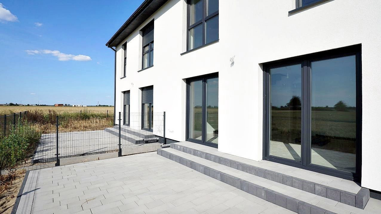 Nowoczesny, dwukondygnacyjny dom z białymi ścianami i ciemnymi oknami, widok z boku na taras wyłożony szarą kostką brukową, z metalowym ogrodzeniem i polami w tle.
