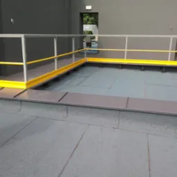 Płaski dach z szarej gumy z żółto-szarą metalową barierką zabezpieczającą, widok z góry na fragment dachu z wyjściami.