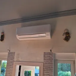 Biały klimatyzator ścienny zamontowany w pomieszczeniu, ozdobiony figurkami aniołków po bokach, widok na okno z zielenią na zewnątrz.