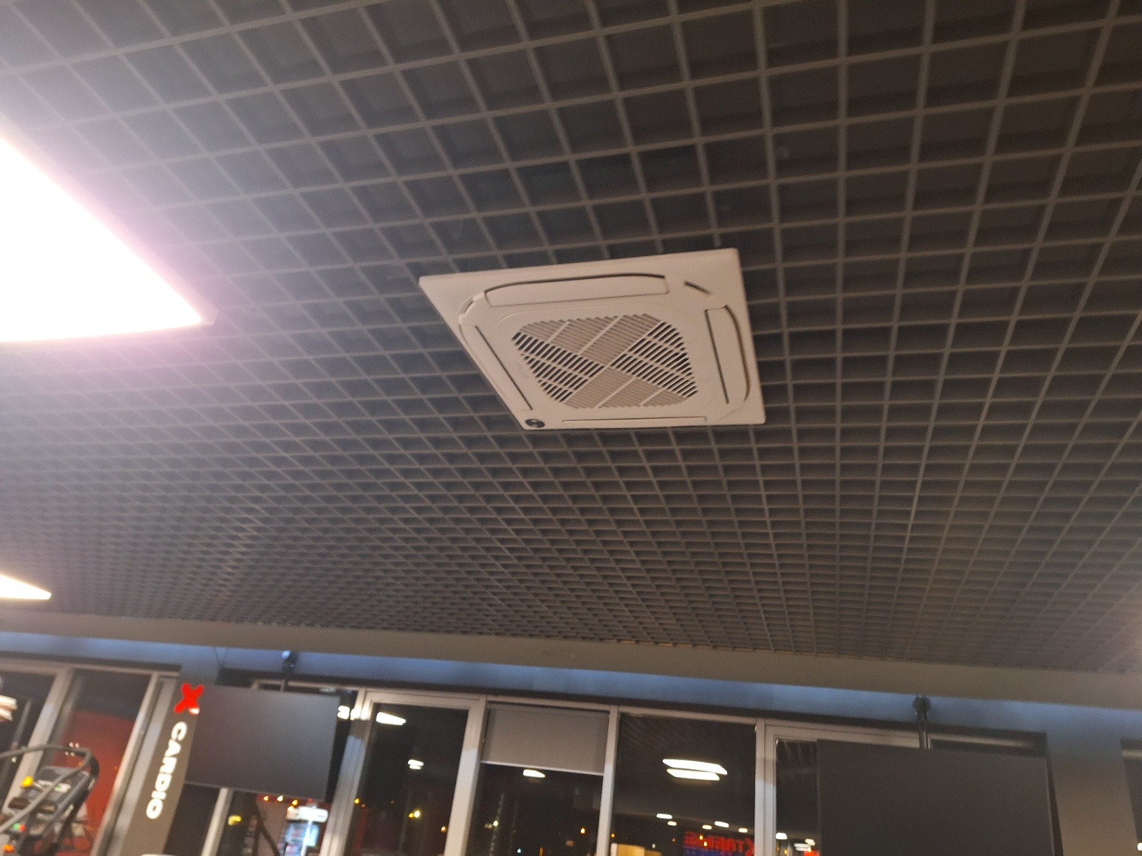 Biała, kasetonowa klimatyzacja sufitowa w rastrowym suficie podwieszanym, widziana pod kątem w jasnym pomieszczeniu biurowym lub usługowym.