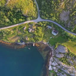 Widok z drona na malowniczą zatokę z kilkoma domami, drogą wijącą się wśr&oacute;d zieleni i skalistego wybrzeża w słoneczny dzień.