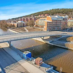 Panorama miasta z rzeką i mostem w słoneczny dzień, widoczne budynki mieszkalne i biurowe na wzgórzu w tle.