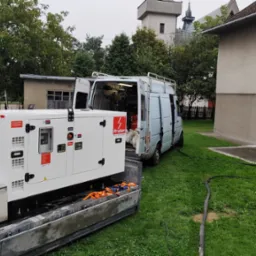 Agregat Proton w busie na trawie, widoczny mężczyzna z napojem, budynki w tle. Sprzęt gotowy do pracy na zewnątrz, zasilanie w trakcie realizacji.