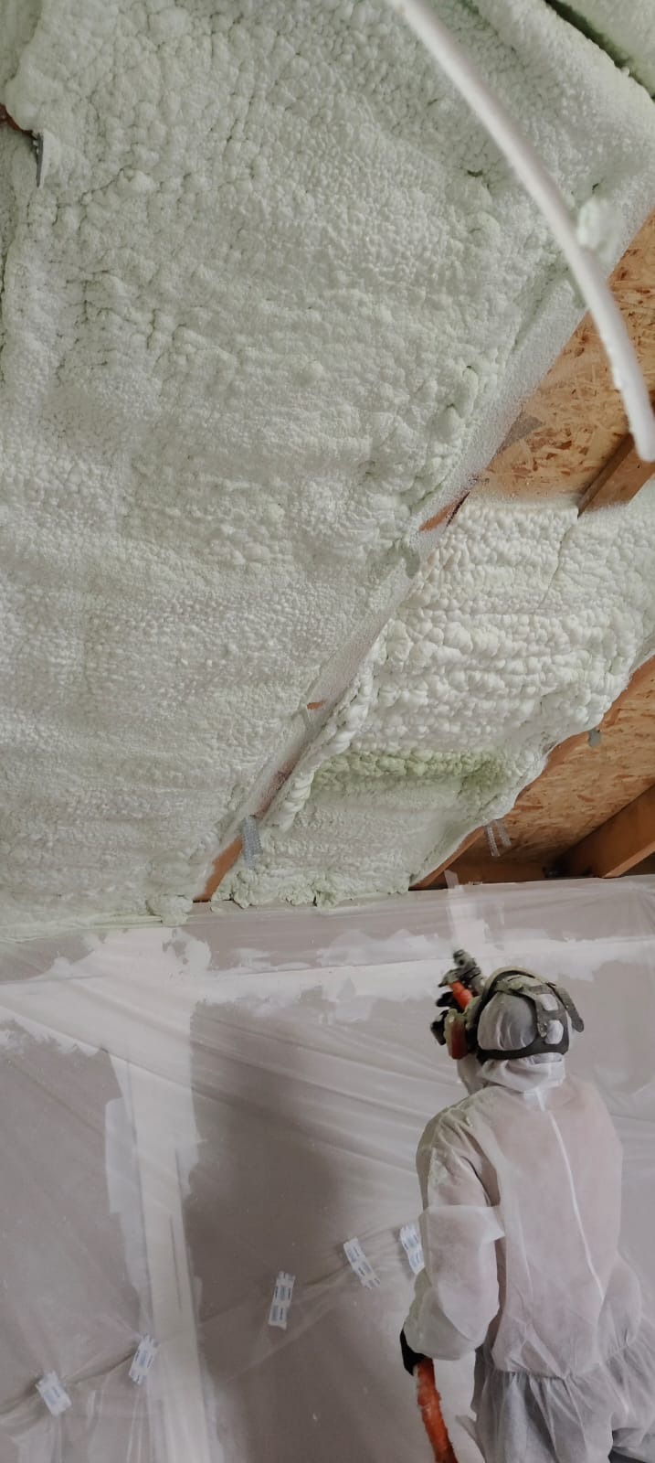Aplikacja pianki PUR na poddaszu przez pracownika w kombinezonie ochronnym. Widoczna struktura piany i elementy konstrukcyjne dachu.
